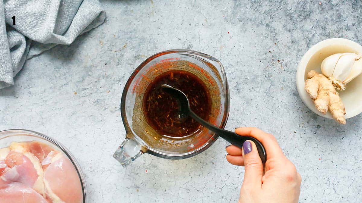 a hand stirring teriyaki sauce in a glass measuring cup with a black spoon. 