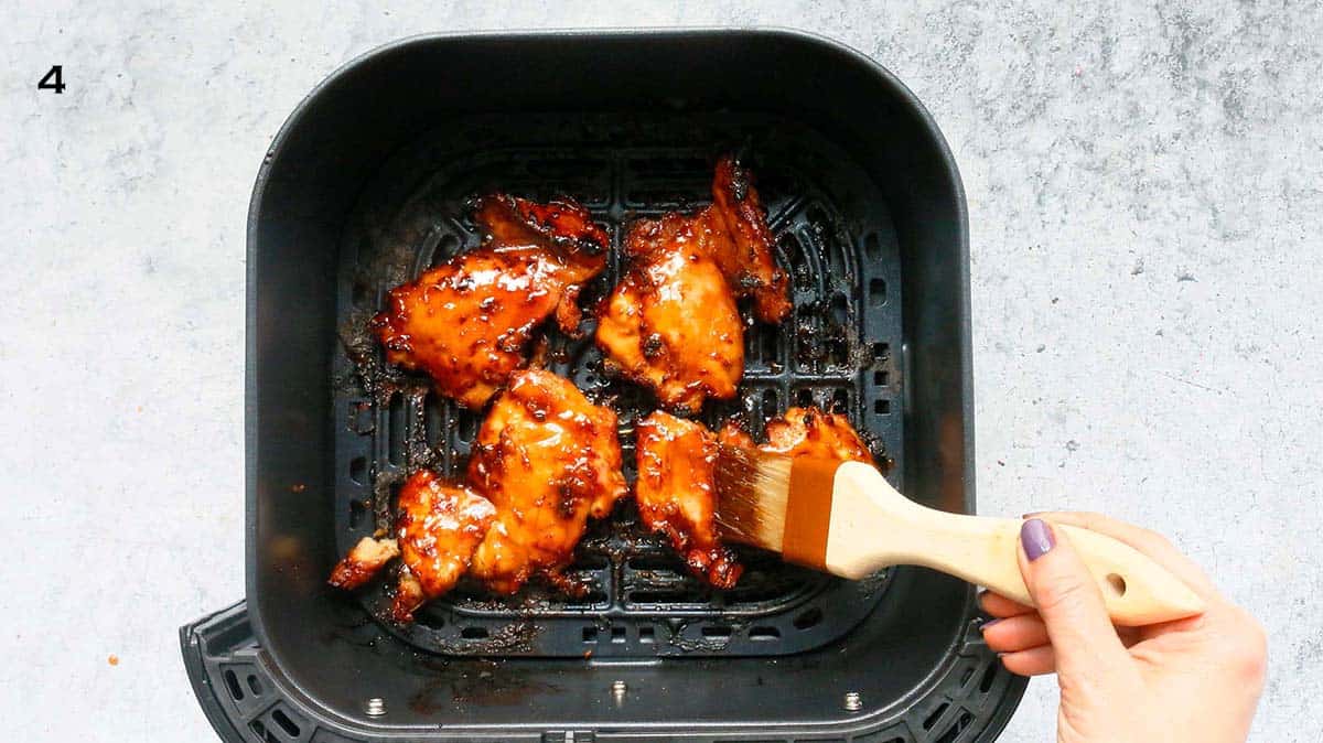 a hand brushing teriyaki sauce on chicken thighs in an air fryer basket.