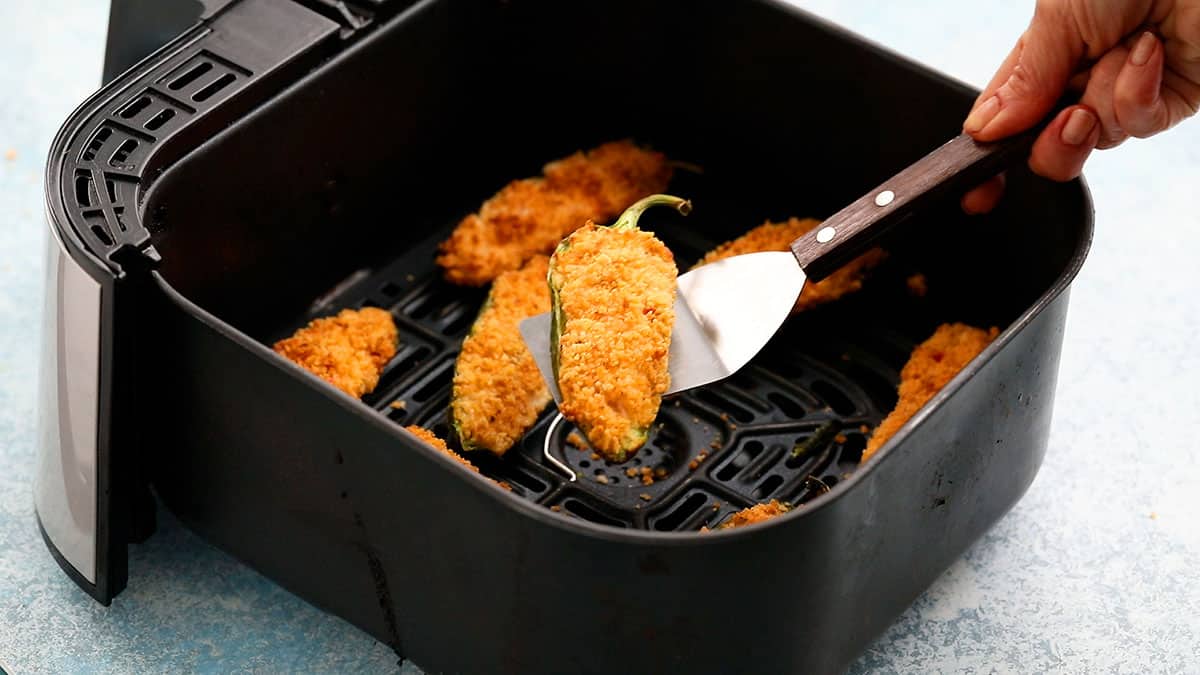a hand lifting one jalapeno popper from an air fryer basket with a flat spatula.