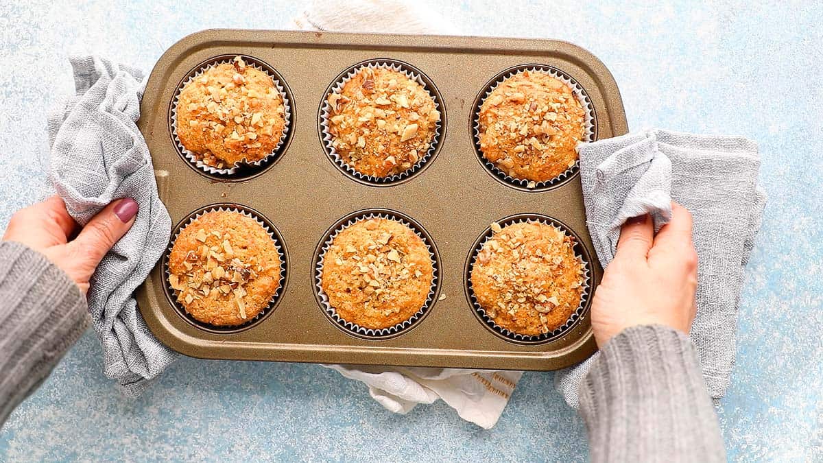 two hands holding a hot baked 6 cup muffin pan with grey kitchen towels.
