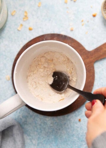 a hand mixing white flour in a white mug using a black spoon. 