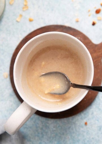 a hand mixing oatmeal cake batter in a white mug using a black spoon.