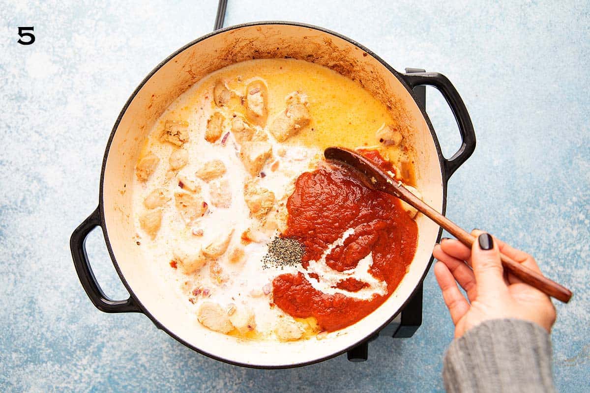 a hand stirring red tomato sauce, heavy cream and stock into a large white skillet.