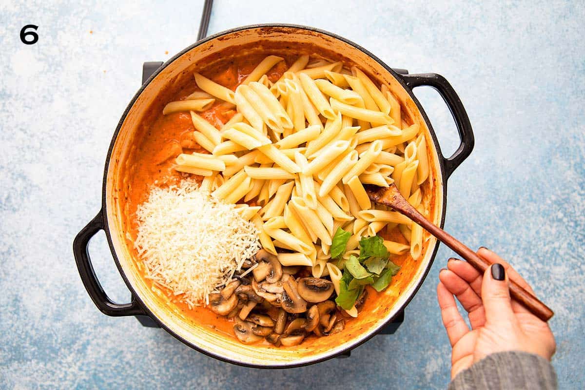 a hand tossing cooked penne pasta, mushrooms, fresh green basil leaves and white cheese in a white skillet.