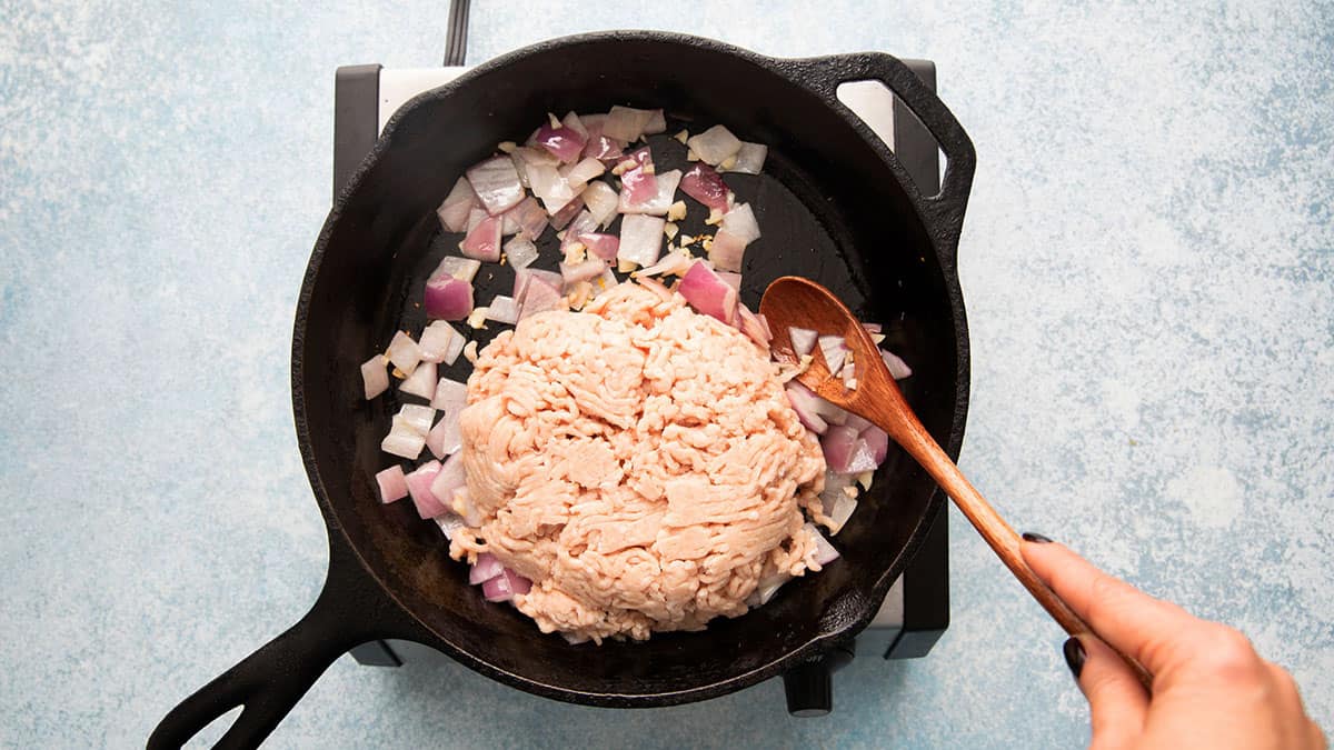 a hand cooking ground chicken and chopped onions in a black skillet.