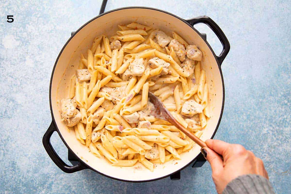 a hand stirring penne pasta and chicken in a large white skillet with wooden spoon.