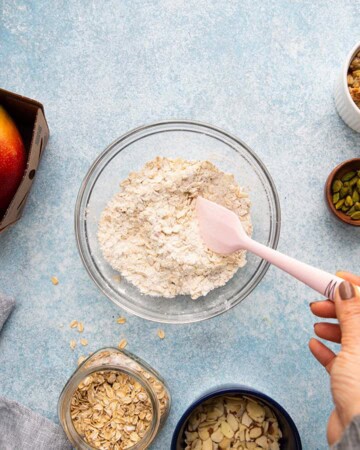 a hand mixing flour and oats in a glass bowl, using a pink spatula.