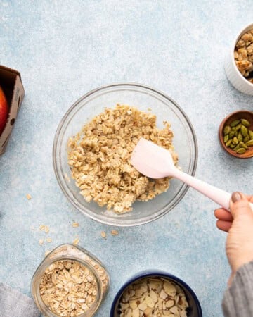 a hand mixing crumble mixture in a glass bowl using a pink spatula.