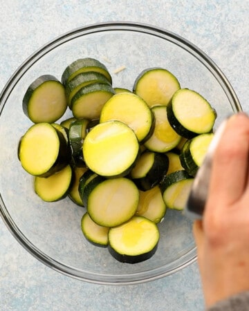 a hand pouring oil over zucchini rounds in a glass bowl.