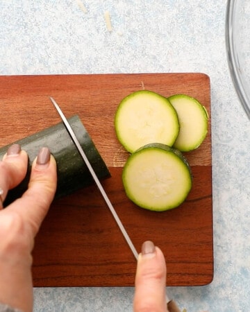 two hands slicing one zucchini into rounds on a wooden board.