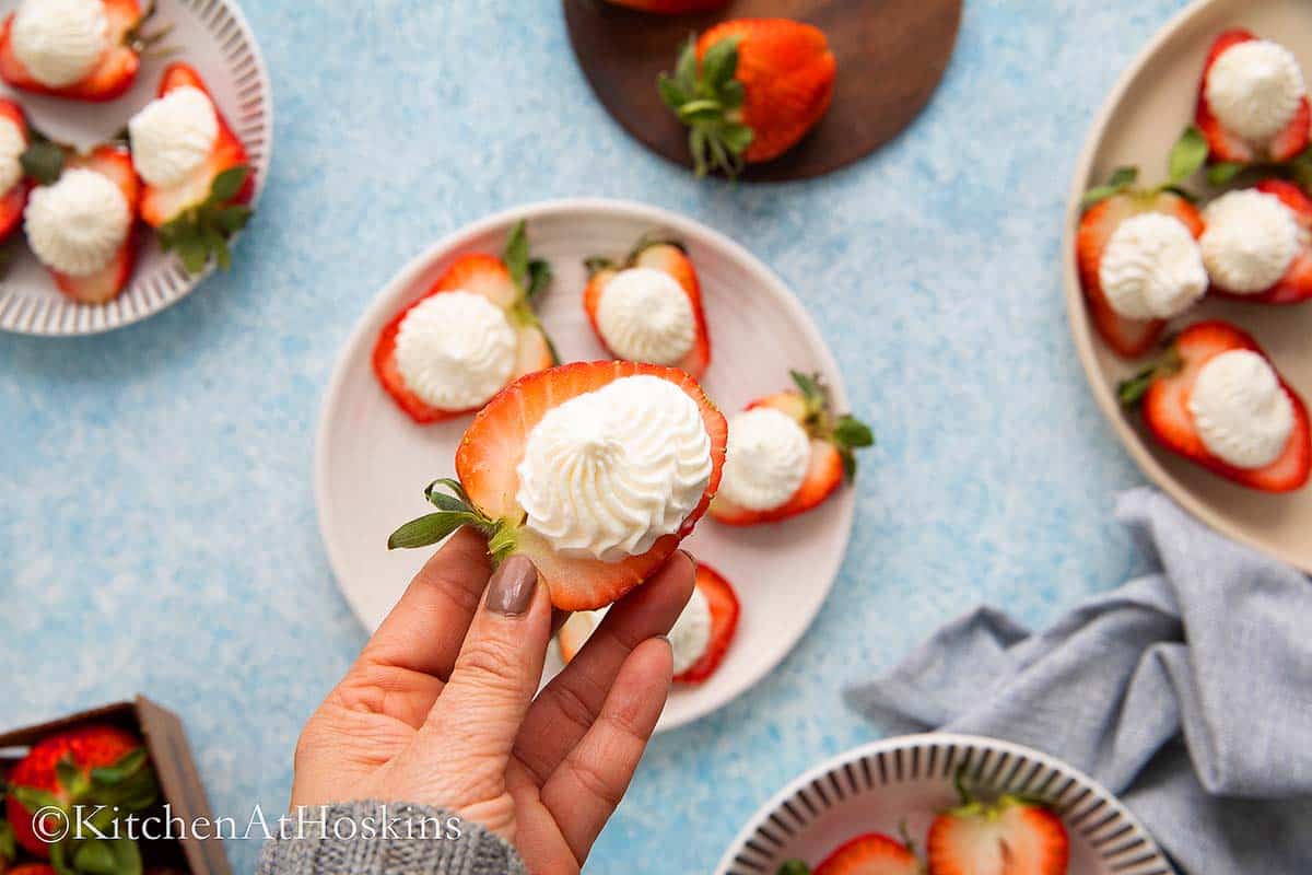 A HAND HOLDING A HALVED STRAWBERRY WITH WHIPPED CREAM.