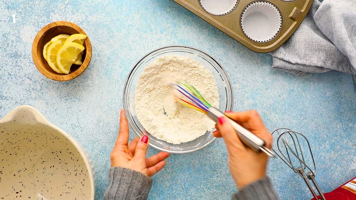 two hands whisking flour in a glass bowl with a whisk.