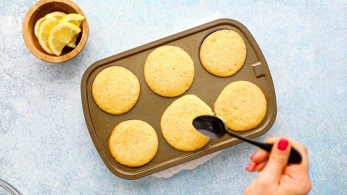 a hand drizzling lemon juice on cupcakes using a black spoon.