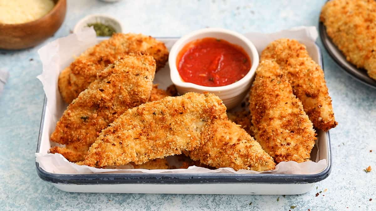 crispy breaded chicken tenders in a white rectangle platter along with tomato sauce.