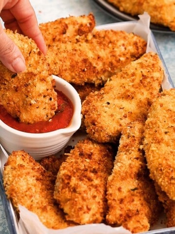 a hand dipping one breaded chicken tender in tomato sauce.