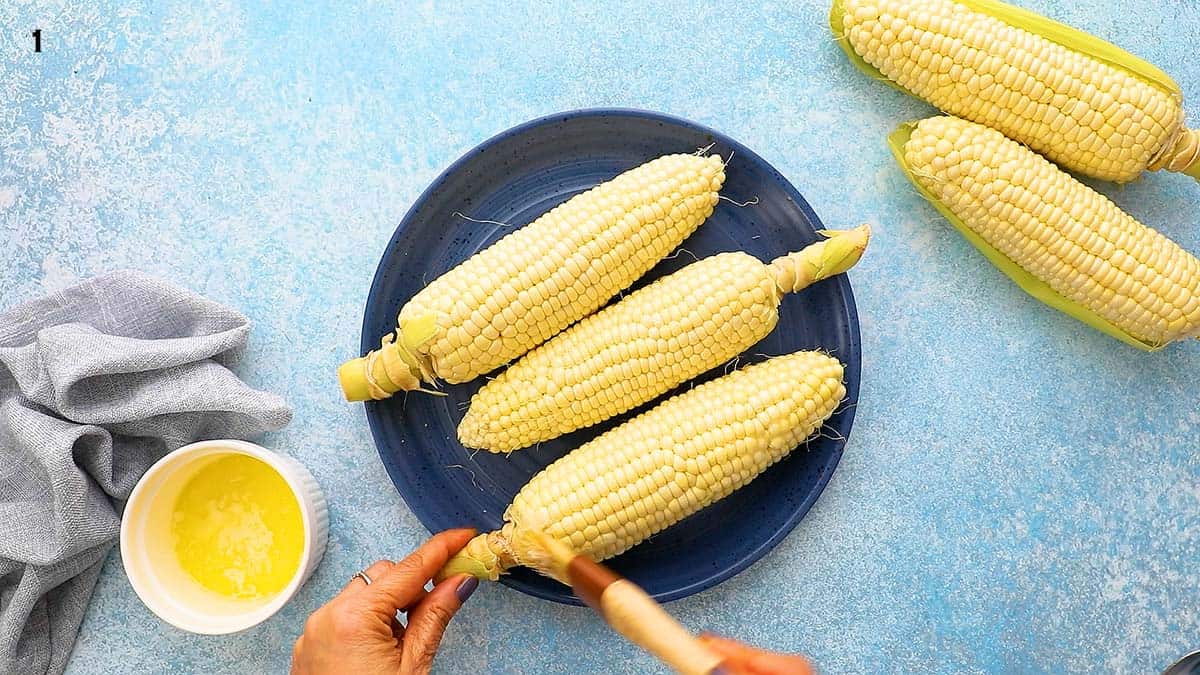 a hand brushing melted butter on 3 corn on the cobs in a blue plate.