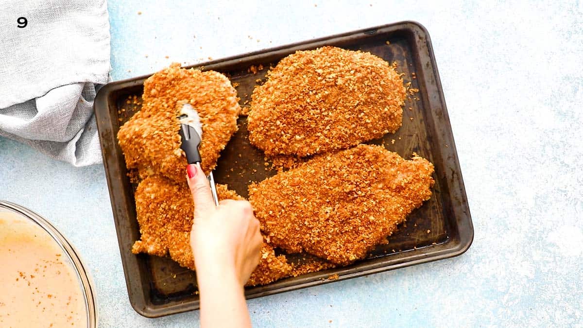 a hand placing a breaded chicken on a black sheet pan using black tongs.