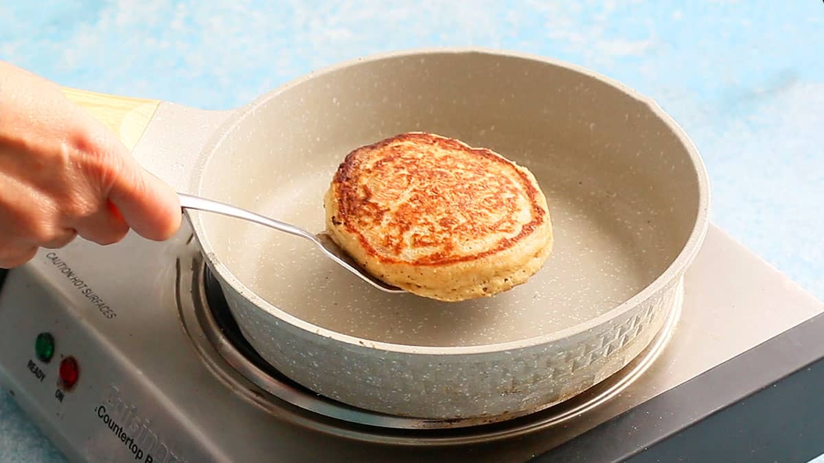 a hand lifting one cooked pancake from a small white skillet.