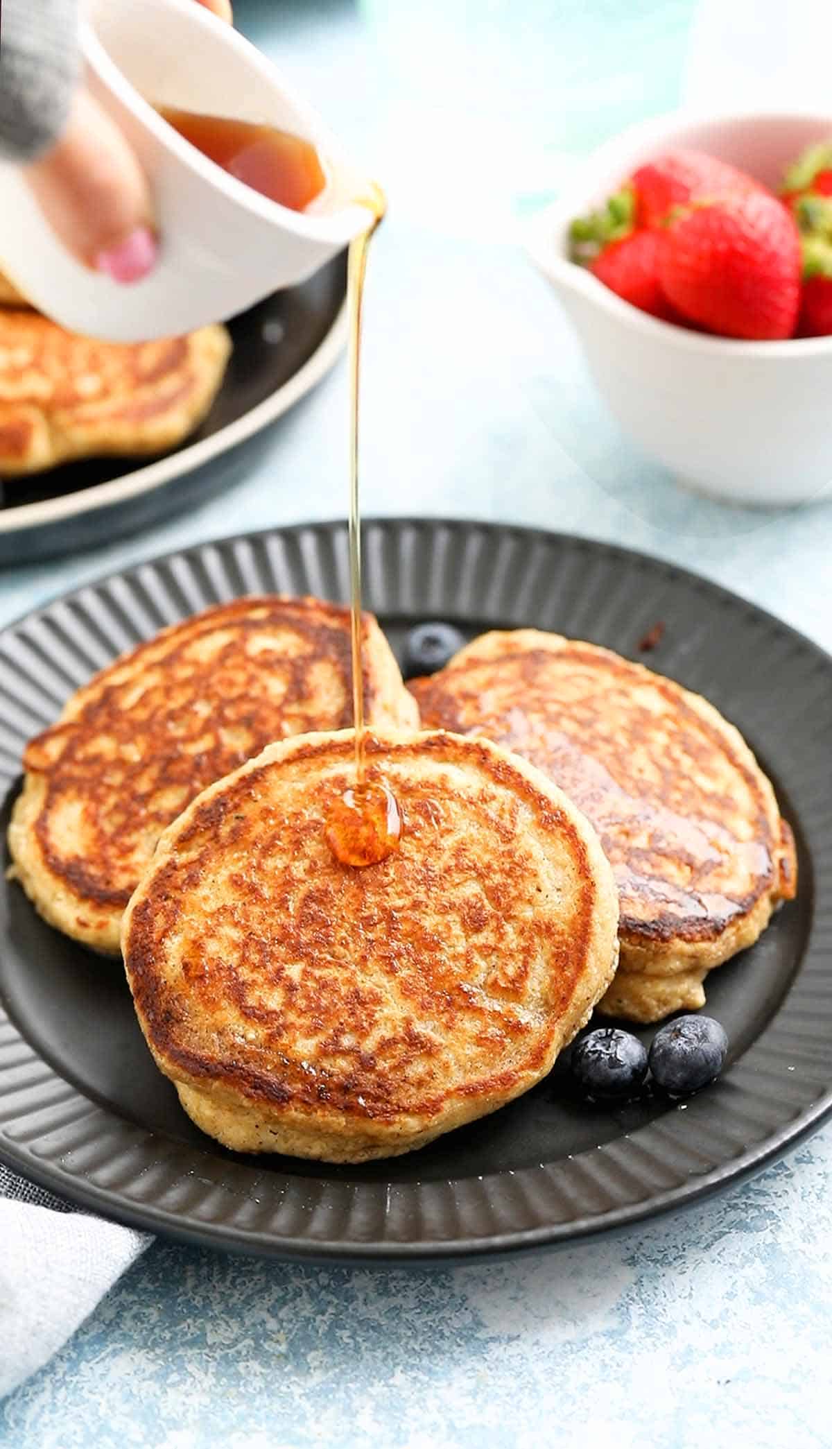 a hand pouring maple syrup over 3 pancakes on a black plate.