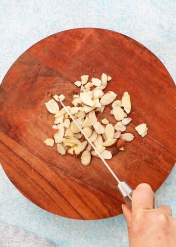 a hand chopping sliced almonds on a round wooden cutting board using a knife.