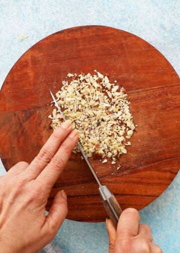 a hand chopping sliced almonds on a round wooden cutting board using a knife.
