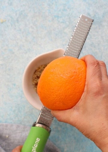 two hands zesting one whole orange using a stainless steel microplane.