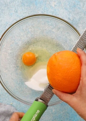 two hands zesting one whole orange using a stainless steel microplane.