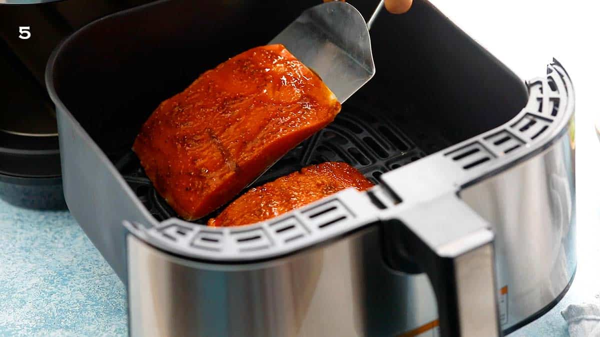 placing one marinated salmon fillet into an air fryer basket using a stainless steel spatula.