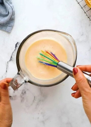 two hands whisking yellow liquid in a glass measuring cup using a colored whisk.