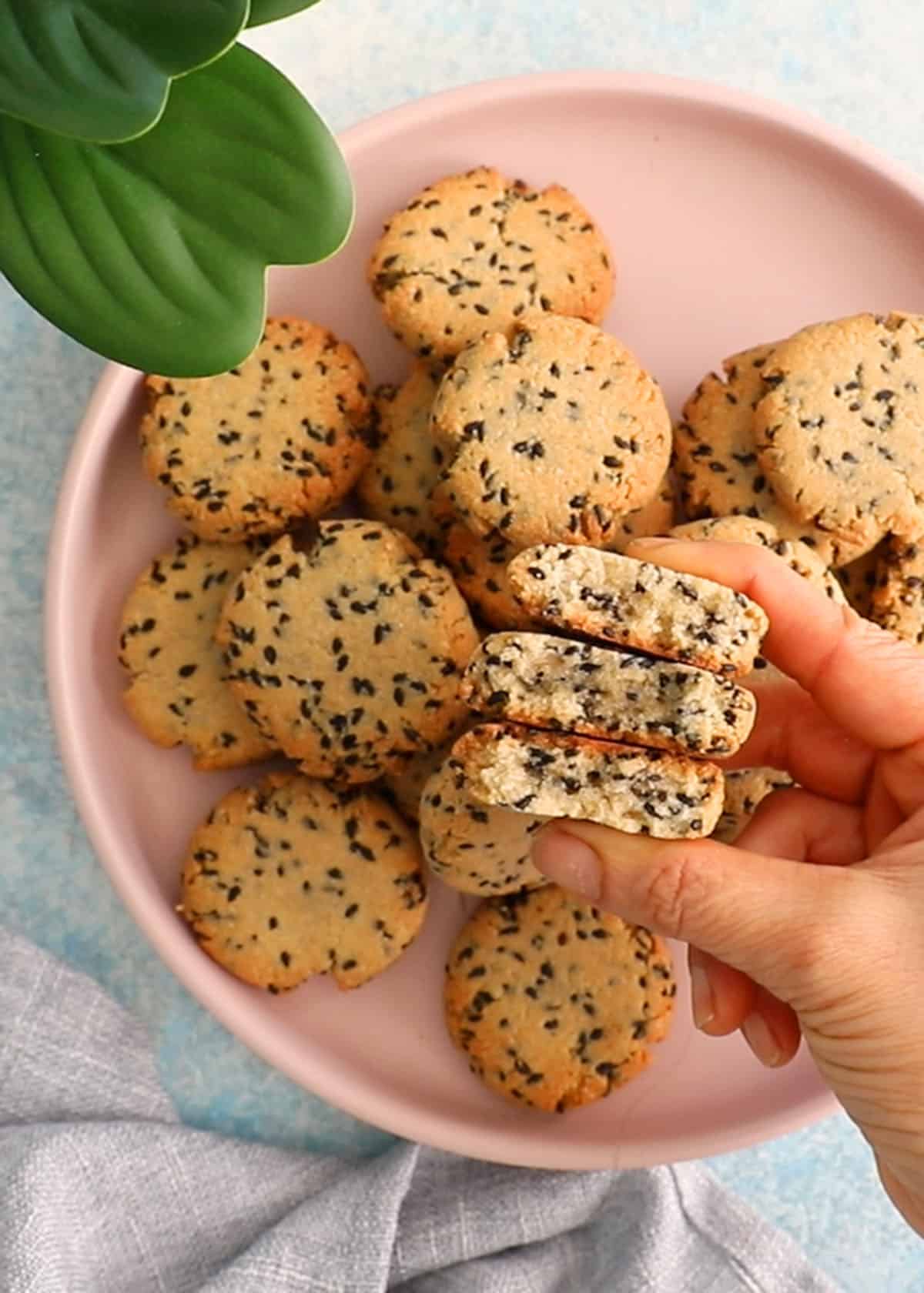 a hand holding balck sesame cookies above a pink plate filled iwth the same.