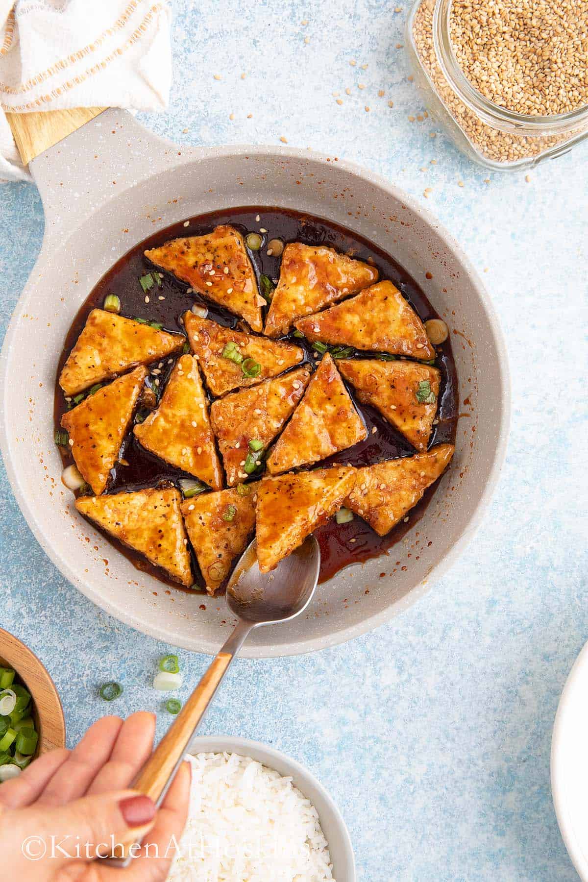 a spoon lifting a cooked tofu from a pan smothered in teriyaki sauce.