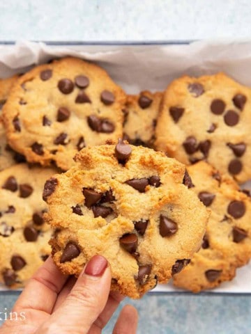 a hand holding one chocolate chip cookie above a platter with the same.