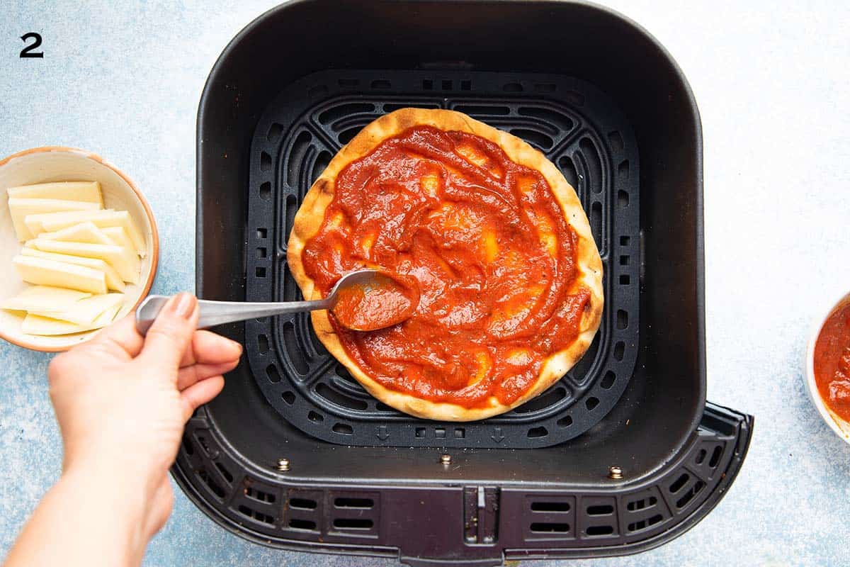 a hand spreading tomato sauce on top of a naan in an air fryer basket.