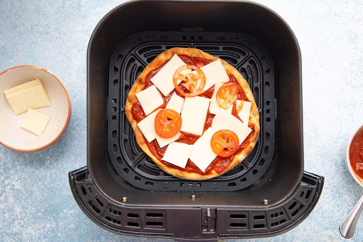 naan bread topped with sliced cheese and tomato in an air fryer basket.