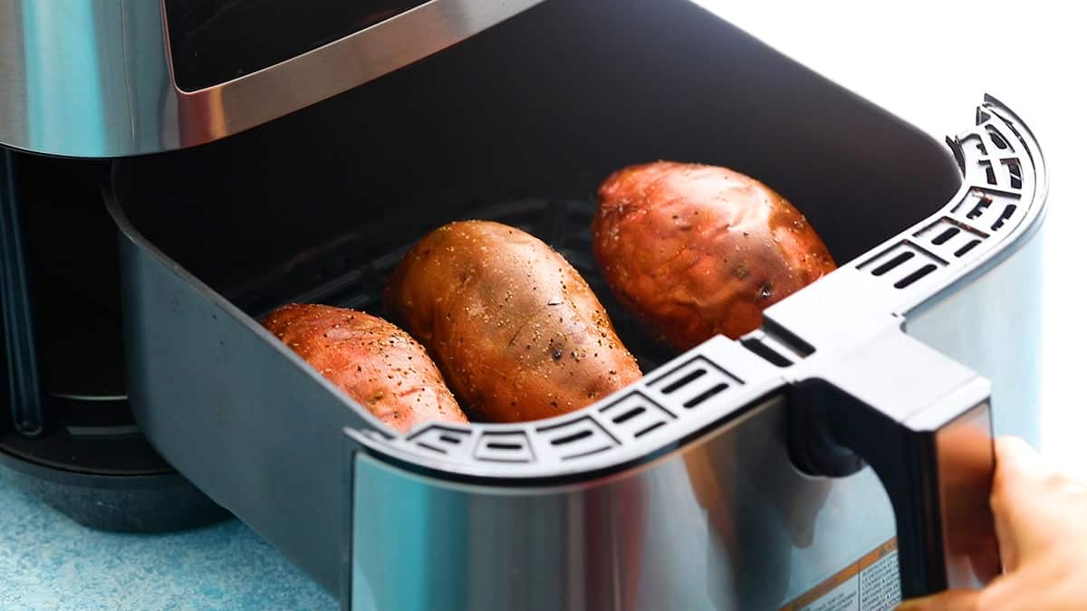 a hand opening an air fryer basket with 3 cooked sweet potatoes.