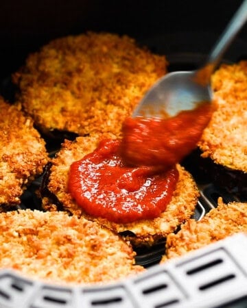 a hand spreading red sauce on top of a breaded eggplant slice.