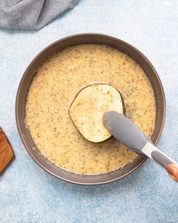 a hand dipping one eggplant slice in wet batter.