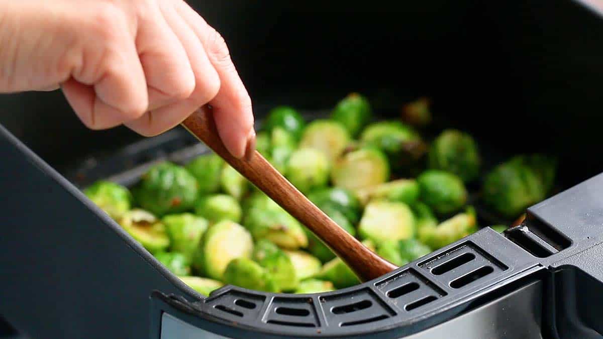 a hand stirring brussels sprouts in an air fryer basket with a wooden spoon. 