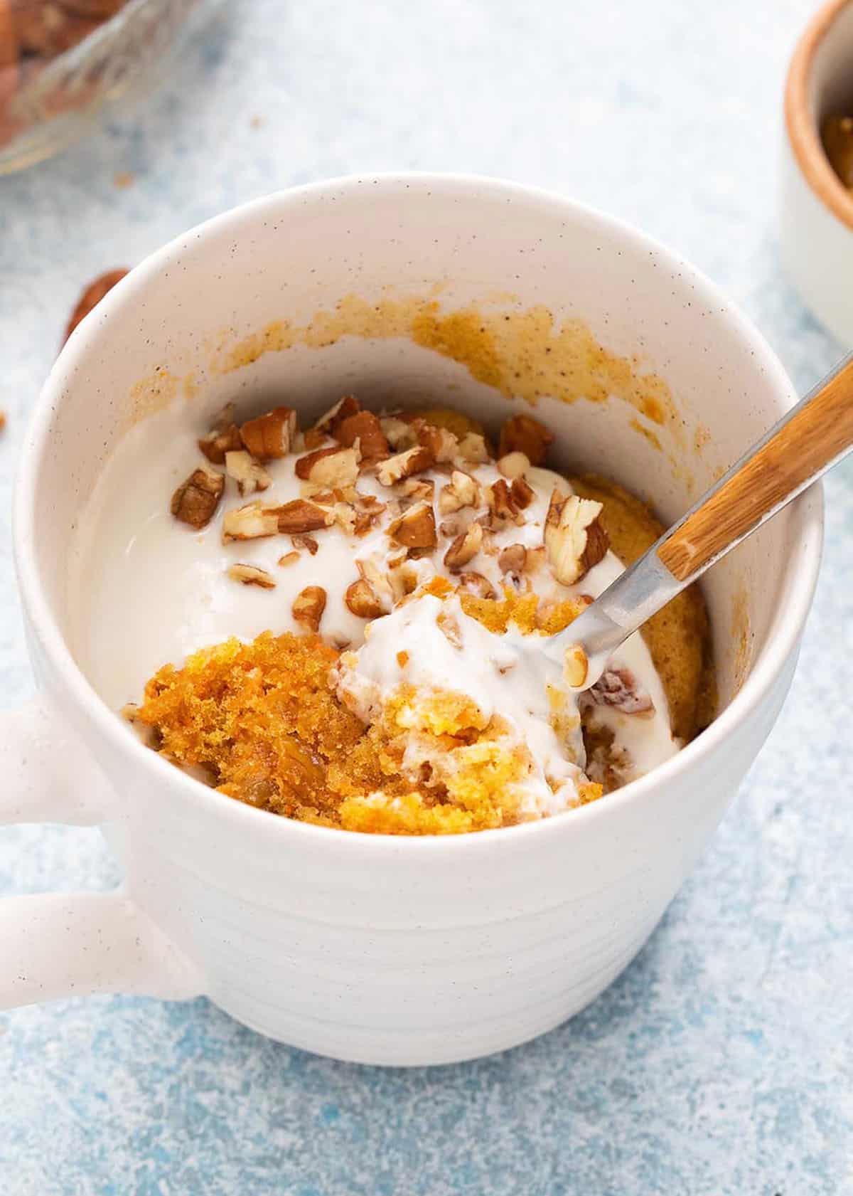 carrot cake topped with white frosting in a white mug along with a spoon.