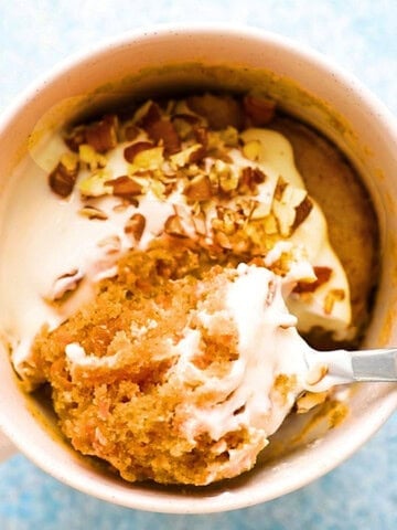 carrot cake topped with white frosting in a white mug along with a spoon.