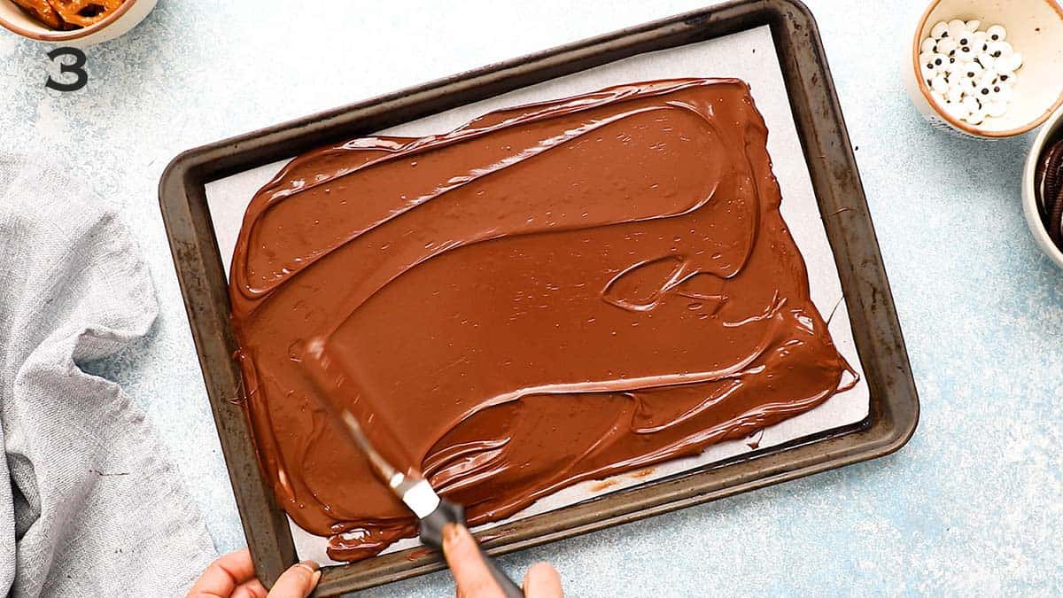 a hand spreading melted chocolate on a parchment lined baking sheet.