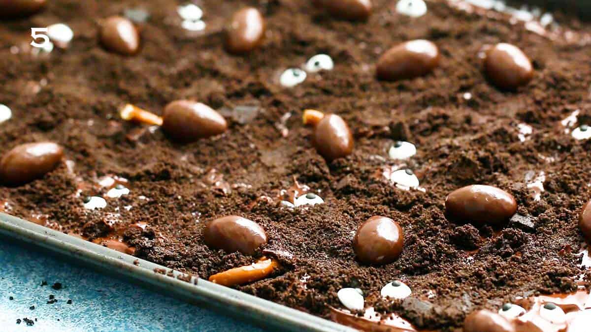 a grey metal baking sheet with crushed oreos, chocolate covered almonds and broken pretzels.