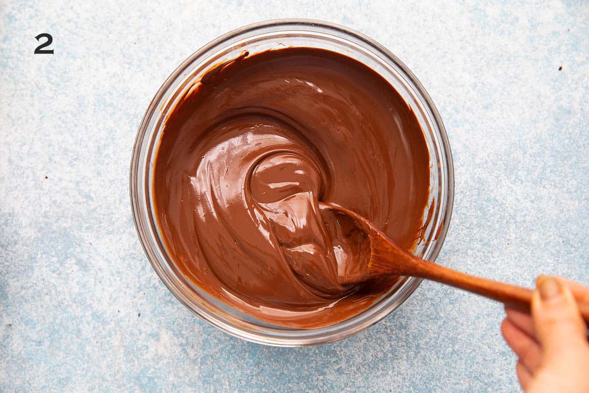 a hand stirring melted chocolate in a glass bowl with a wooden spoon.