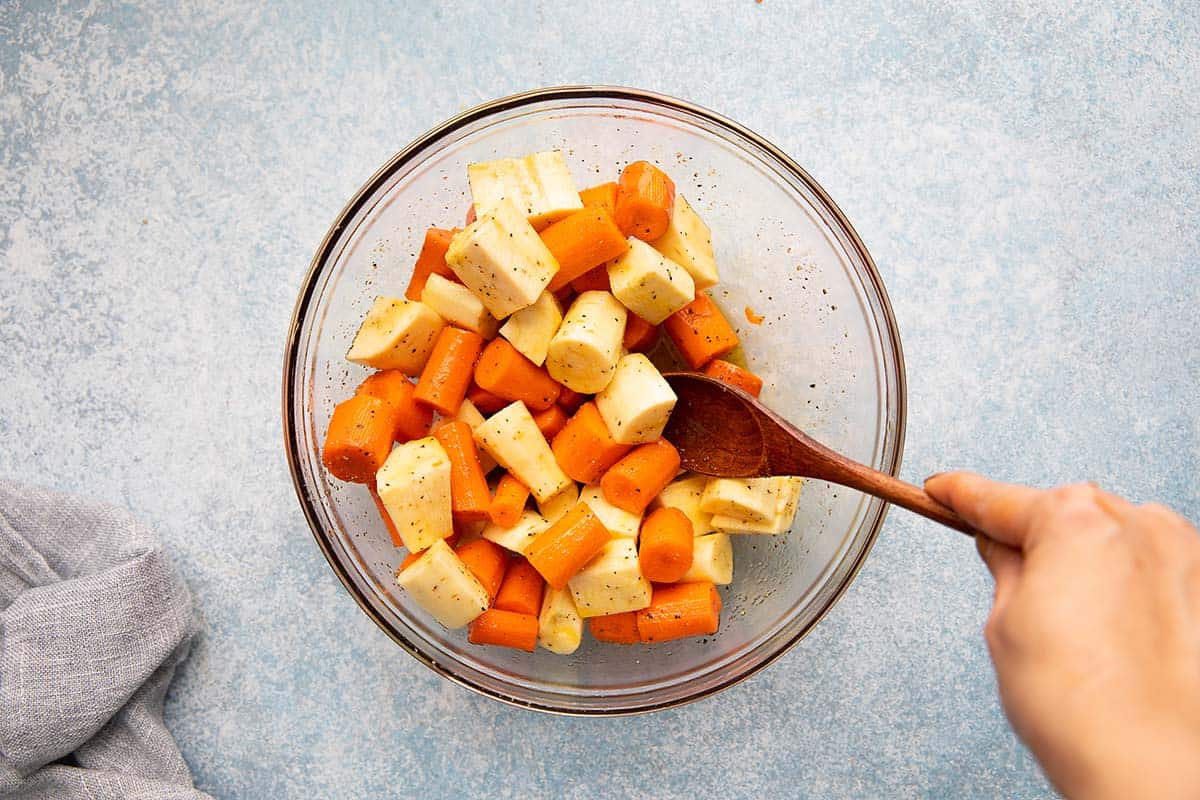 tossing cut carrots and parsnips with seasoning in a glass bowl.