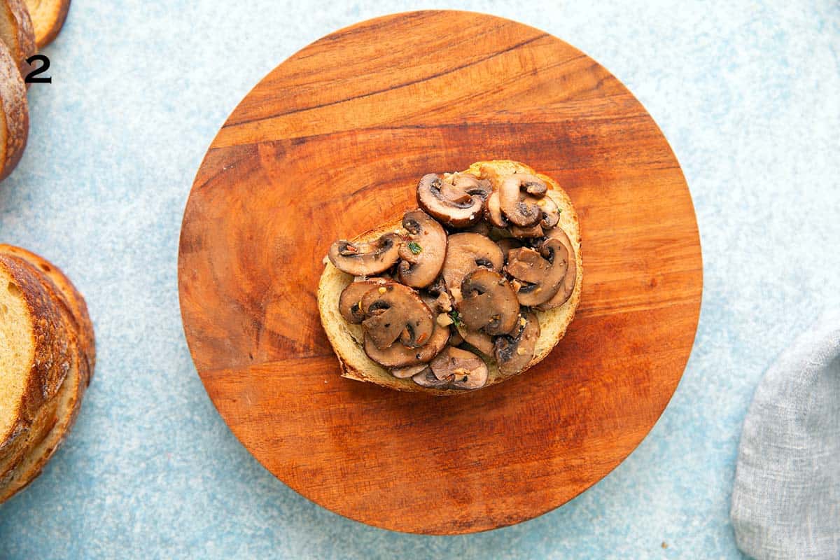 slice of bread placed on around wooden board and topped with cooked mushrooms.