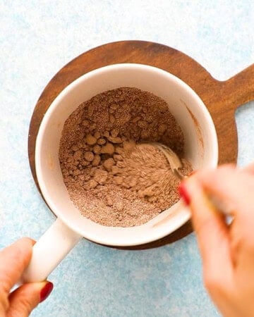 two hands mixing brown flour in a white mug.