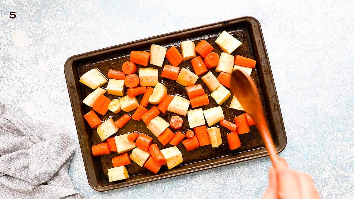 a hand tossing chopped carrots and parsnips in a metal baking sheet.