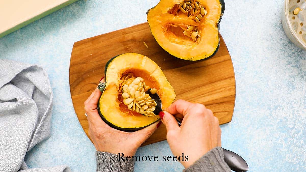 two hands scooping out the seeds from an acorn squash halve using a spoon.