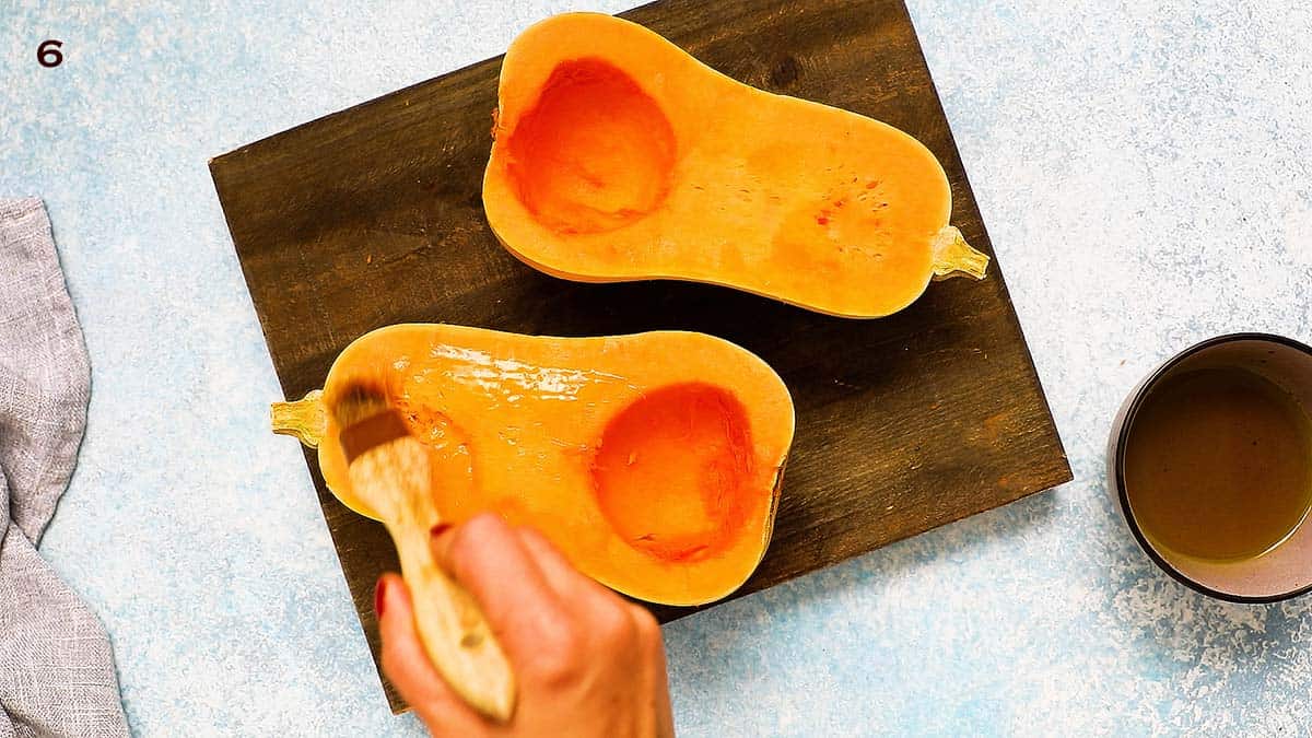 a hand brushing butternut squash halves with oil, using a pastry brush.