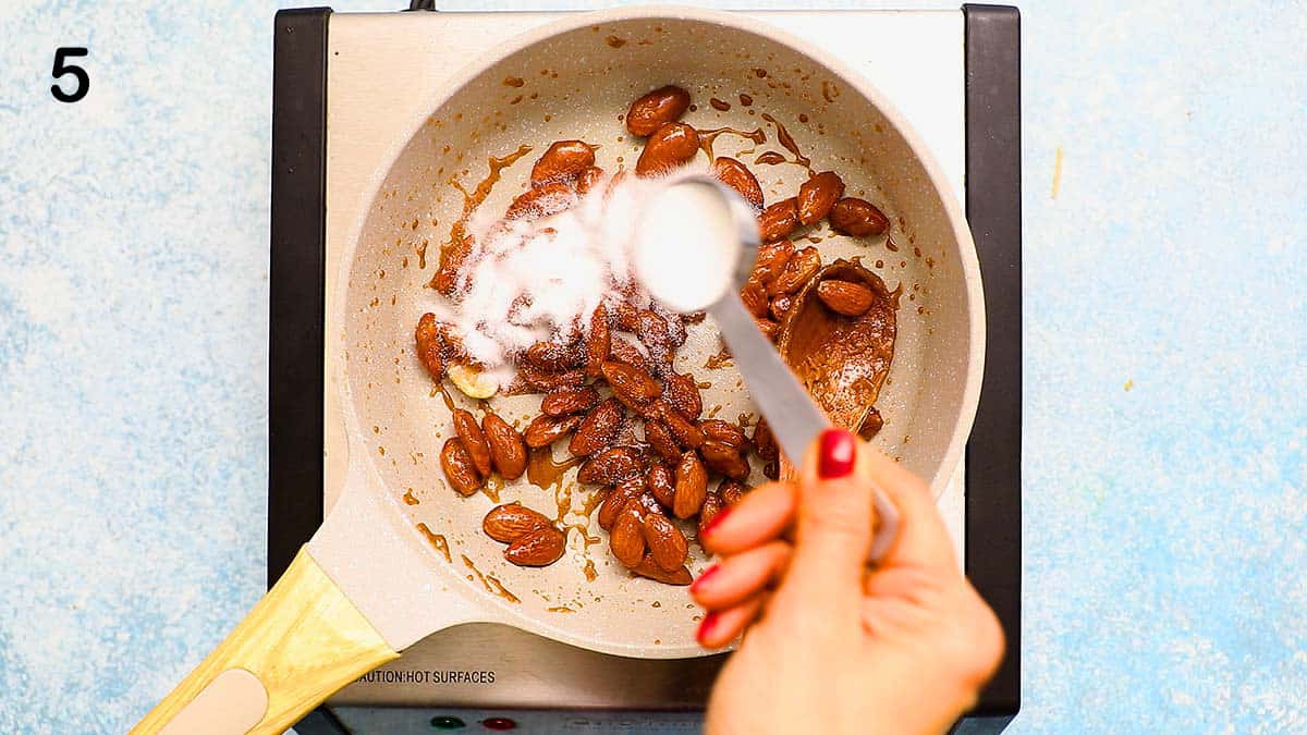 a hand sprinkling sugar over candied almonds in a white skillet.